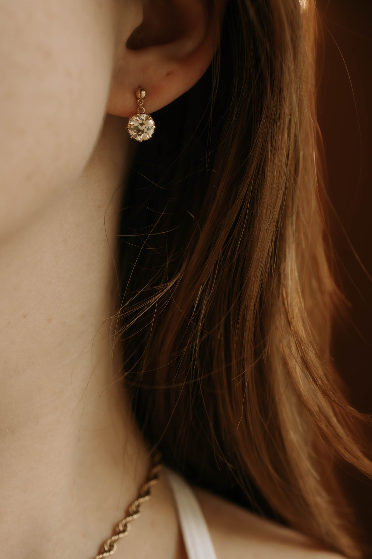 A close-up lifestyle shot of a model wearing a round diamond drop earring The warm, golden-hour lighting highlights the delicate jewelry against the model's red hair and skin.