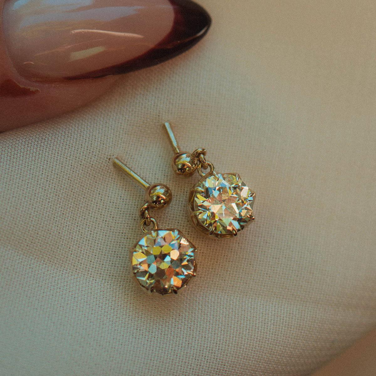 A pair of gold-tone drop earrings with round, old european warm diamonds resting on a soft beige fabric surface, with a hand featuring a dark French-tipped manicure visible in the upper left corner.