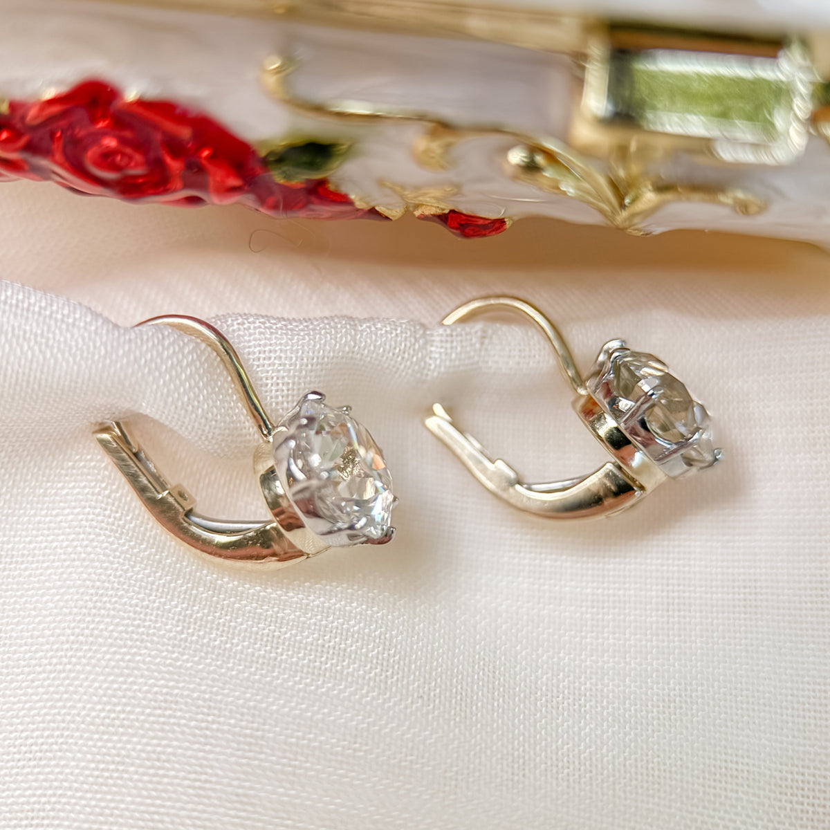 A pair of gold-toned leverback earrings with round old european cut diamonds displayed on a textured white fabric surface with a decorative red and gold clutch bag in the background.