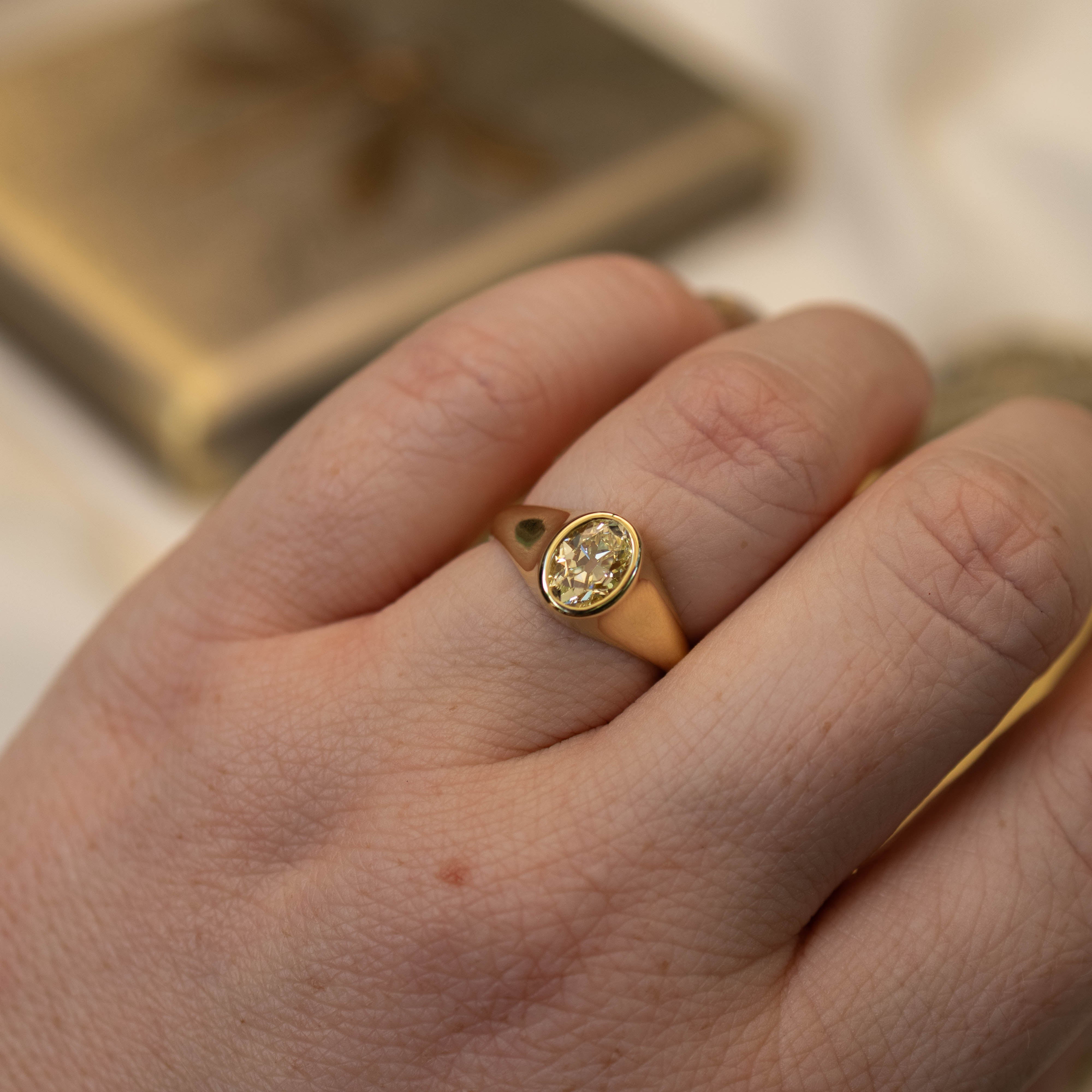 A close-up of a person's hand wearing a gold signet ring with an oval yellow diamond gemstone, set against a soft, out-of-focus background.