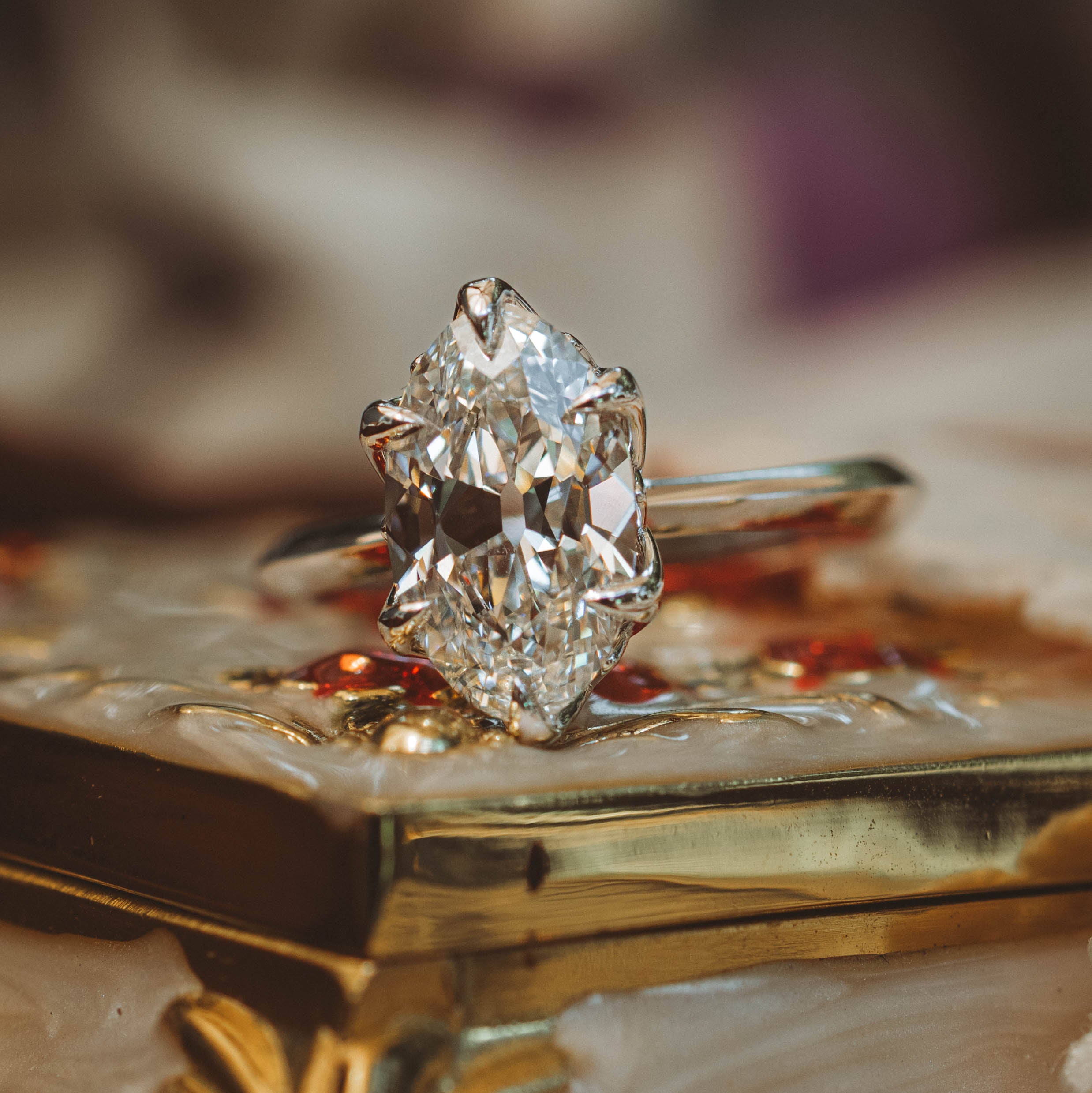 A close-up of a diamond ring with a an oval center stone and a white gold band, resting on an ornate gold and cream decorative box.