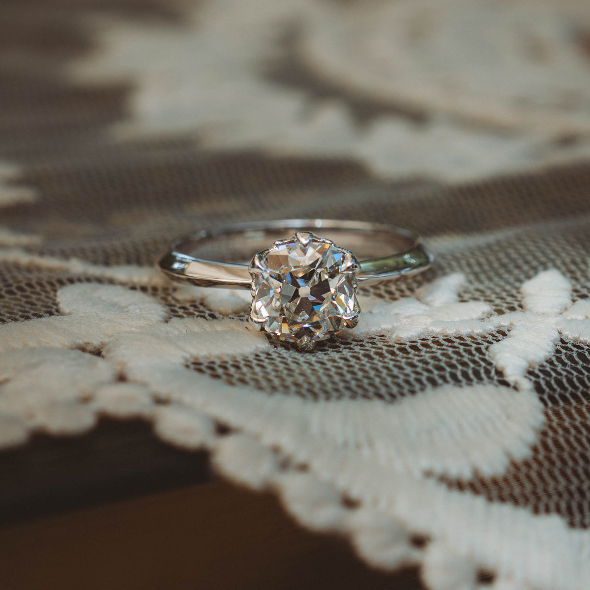 A silver engagement ring with a round brilliant-cut diamond sits on a lace-covered surface.