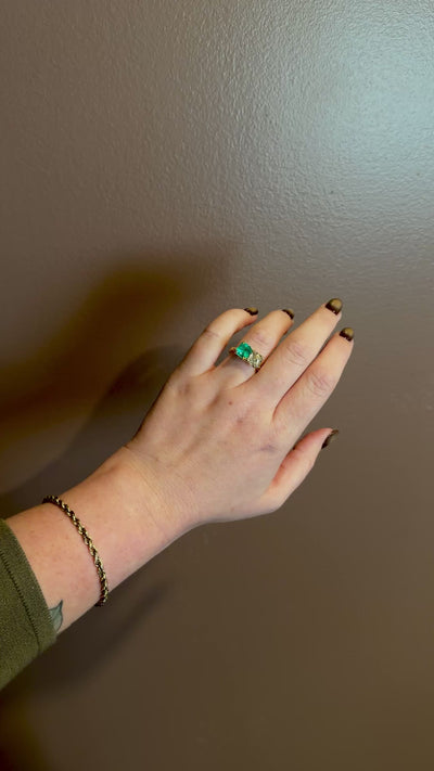 hand moving in front of brown wall wearing yellow gold ring with green square emerald and yellow cushion diamond side by side 