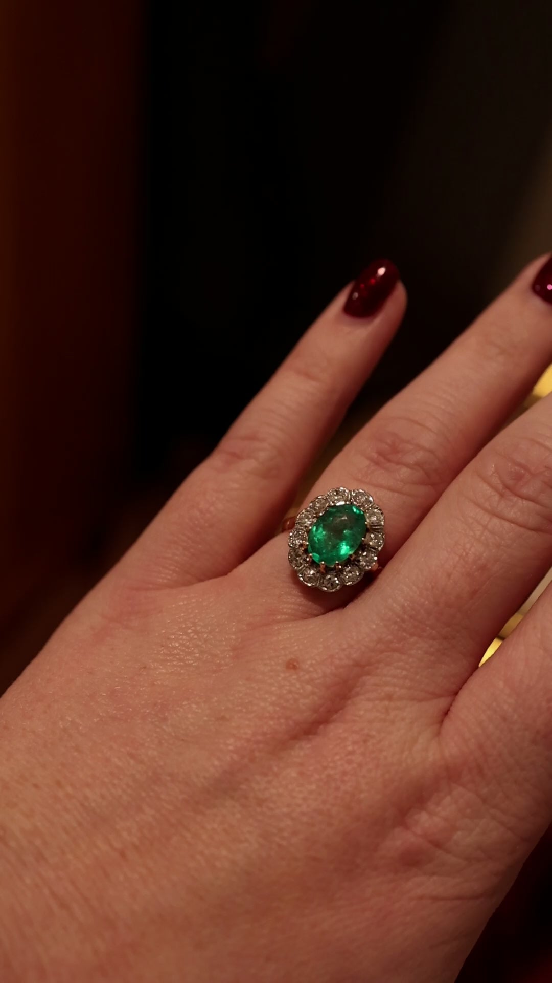green emerald gemstone ring surrounded bu diamond halo worn on hand with dark brown and red background.