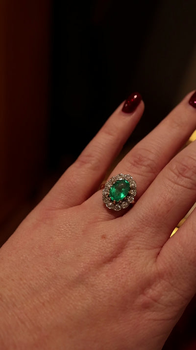 green emerald gemstone ring surrounded bu diamond halo worn on hand with dark brown and red background.