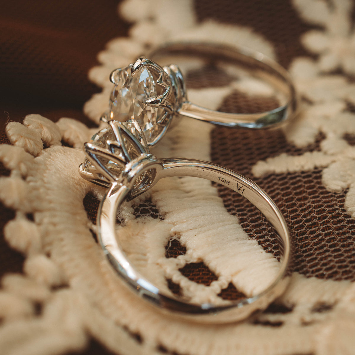 Two silver rings, including a prominent engagement-style ring with a large clear gemstone, resting on a cream lace textile over a dark brown surface.