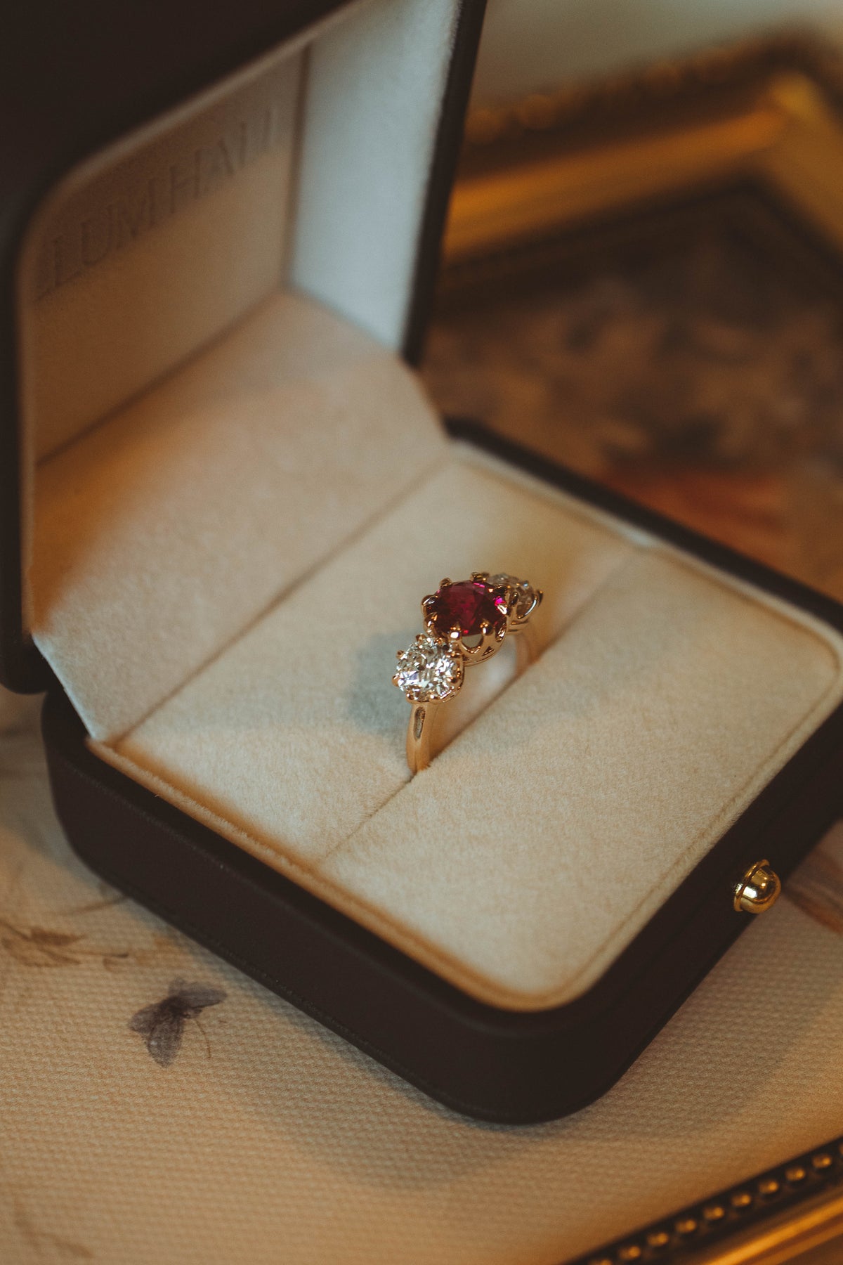 ruby and diamond three stone engagement ring set in rose gold placed in jewelry box sitting on top of painting. 