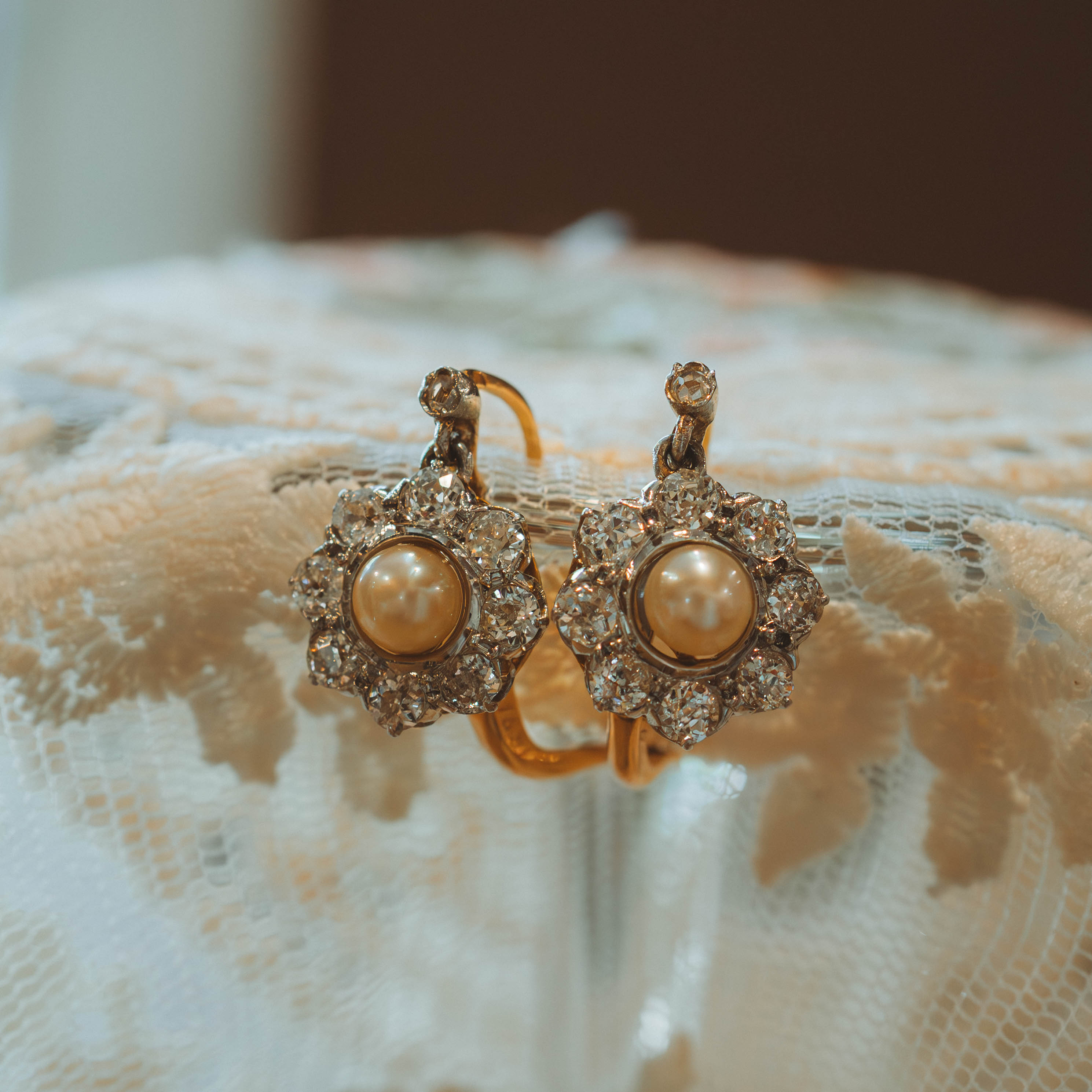Pair of gold earrings with pearl centers & diamond halo on a textured lace surface