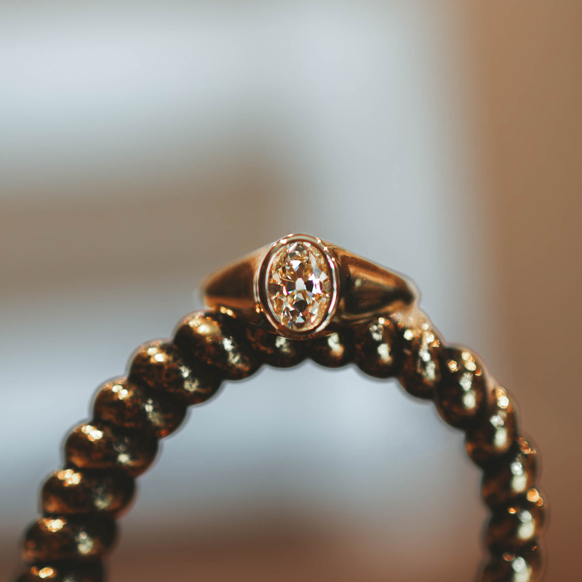 A gold ring with an antique oval yellow diamond displayed on a dark beaded stand against a warm, blurred background.