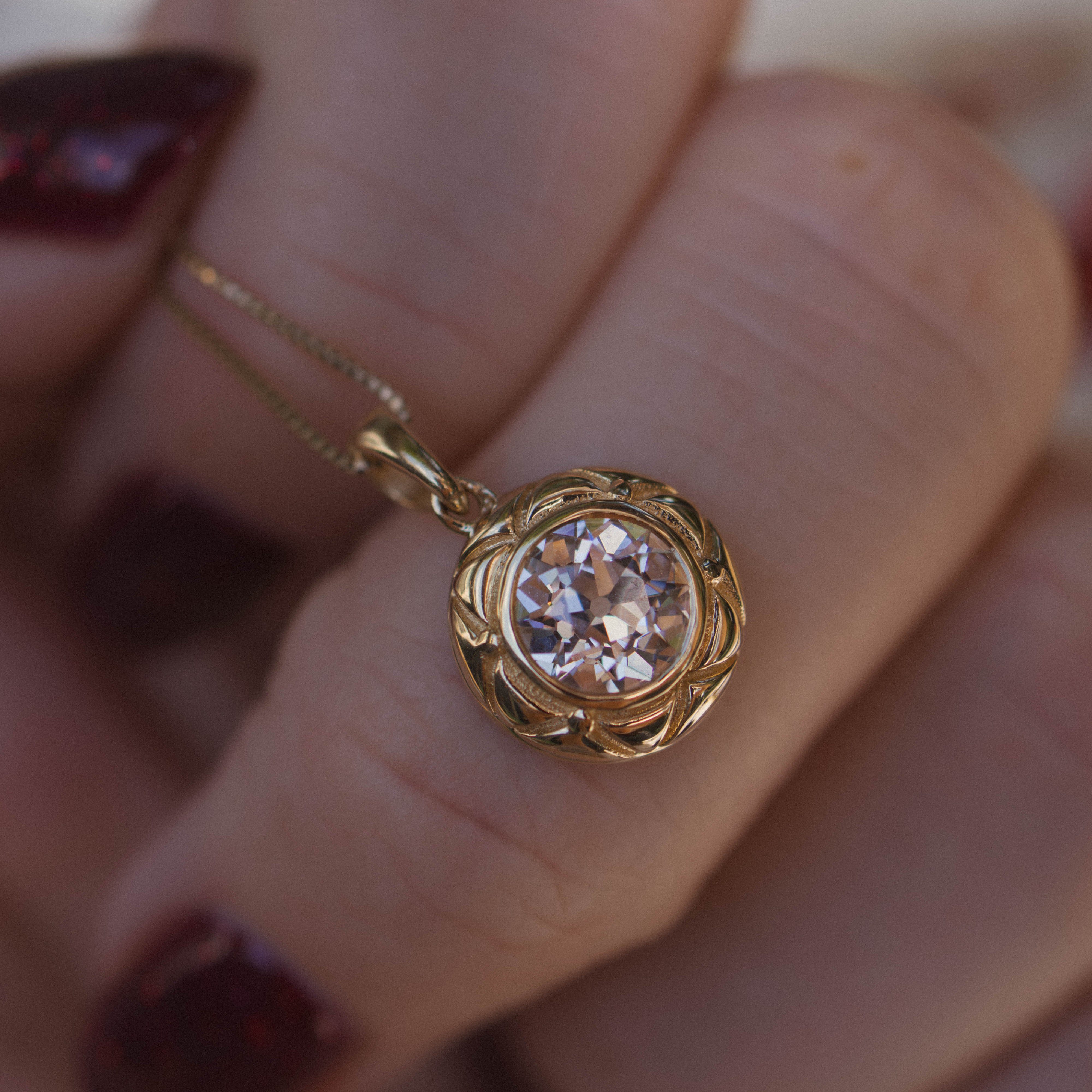 A gold pendant necklace with a clear round old european diamond held on a person's finger. The pendant has a textured, yellow gold scalloped border around the diamond