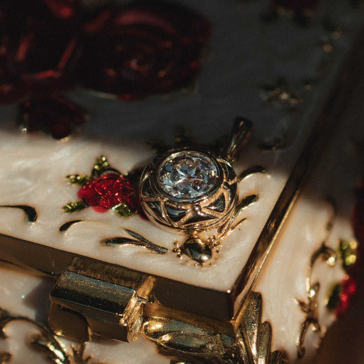 A close-up of an ornate gold and white enamel jewelry box decorated with red roses, featuring a small clear gemstone pendant resting on its lid.