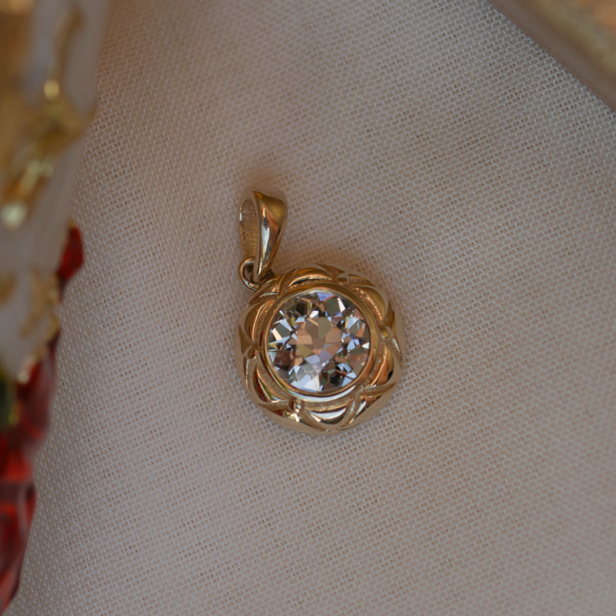 A gold pendant with a clear rround old european diamond gemstone, resting on a textured beige fabric surface.