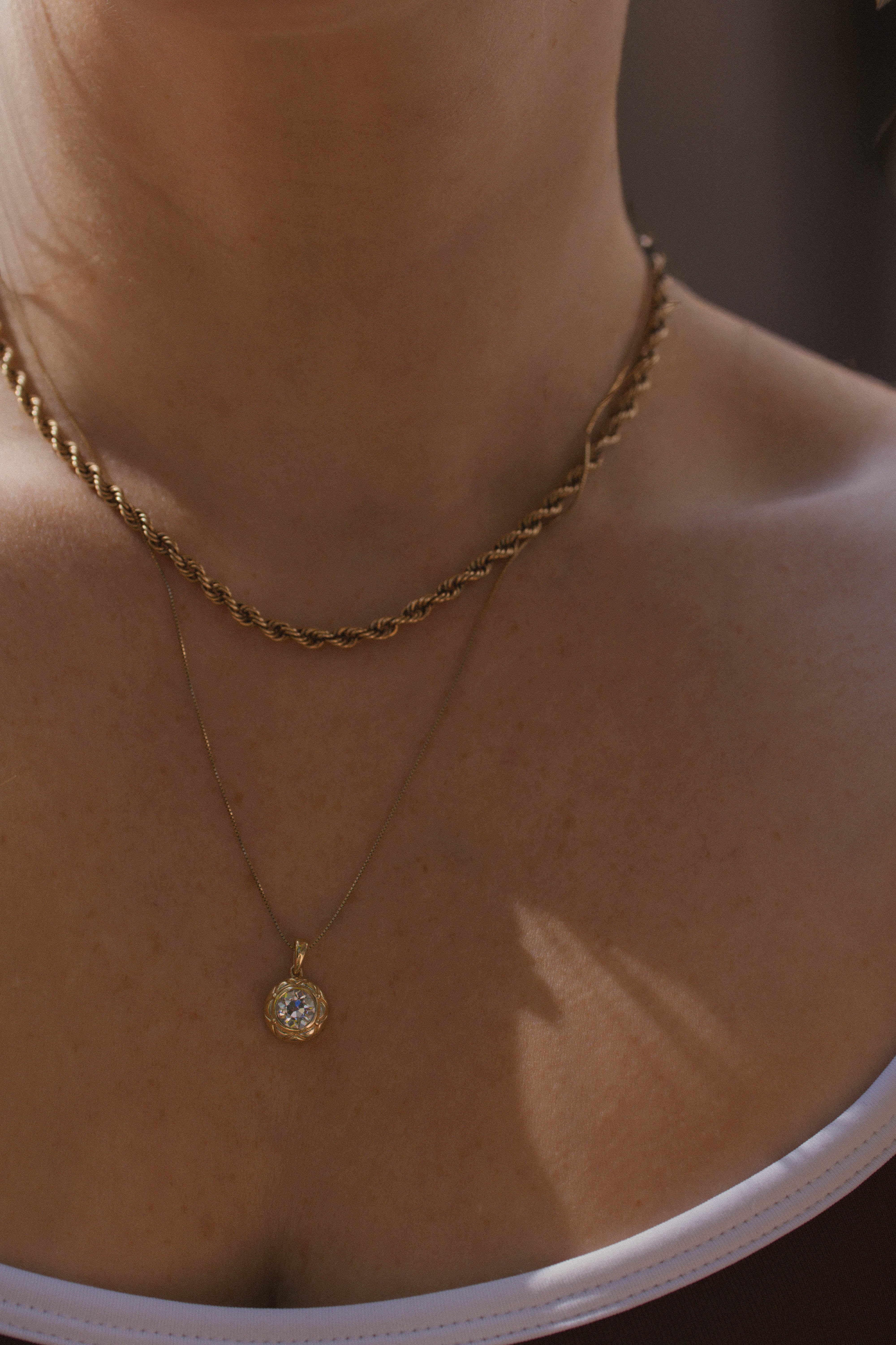 A close-up of a person's neck and upper chest wearing layered gold necklaces, including a twisted chain and a delicate chain with a small round pendant.