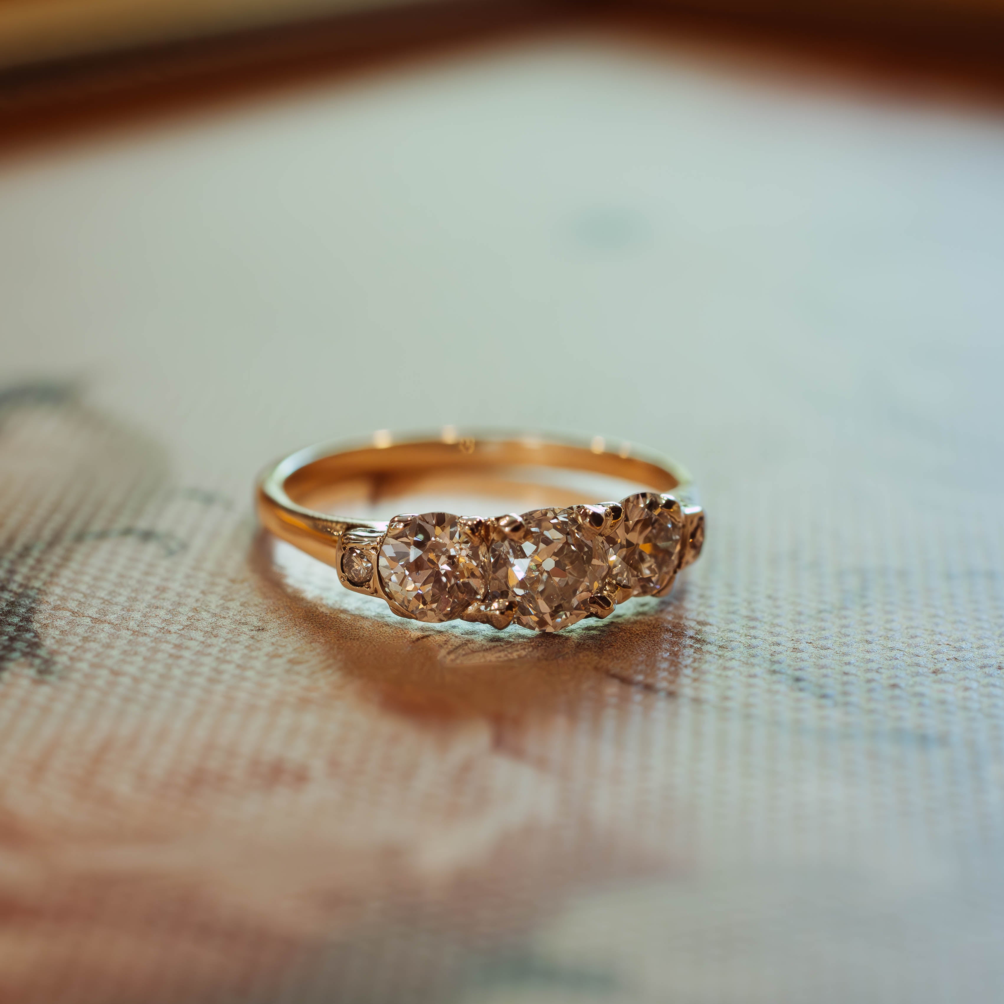 A gold ring with three small round diamonds set in a row, resting on a textured beige surface with a soft-focus background.