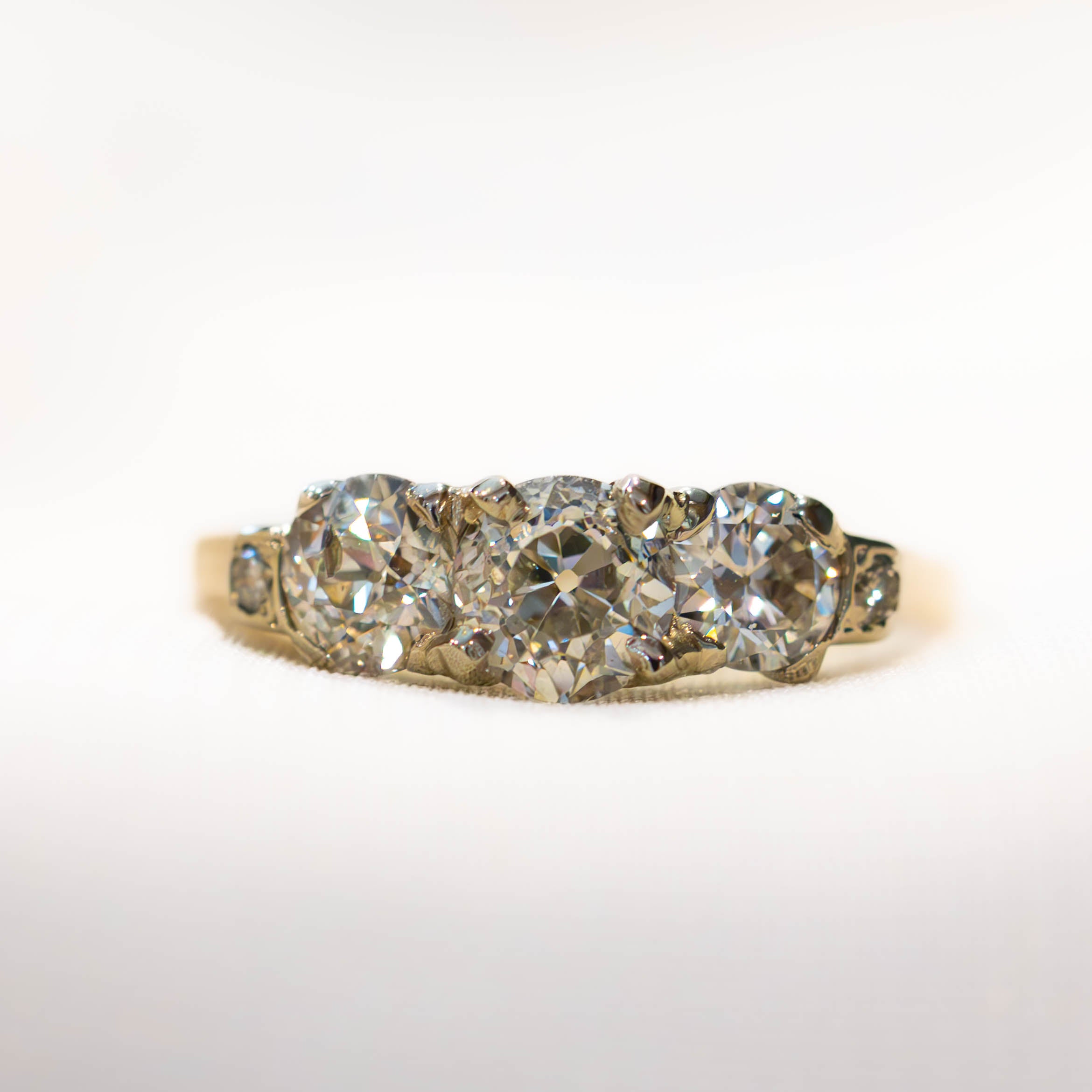 A vintage-style three-stone diamond ring with a yellow gold band, featuring three round brilliant-cut diamonds in a row with smaller accent diamonds on the shoulders, set against a plain white background.