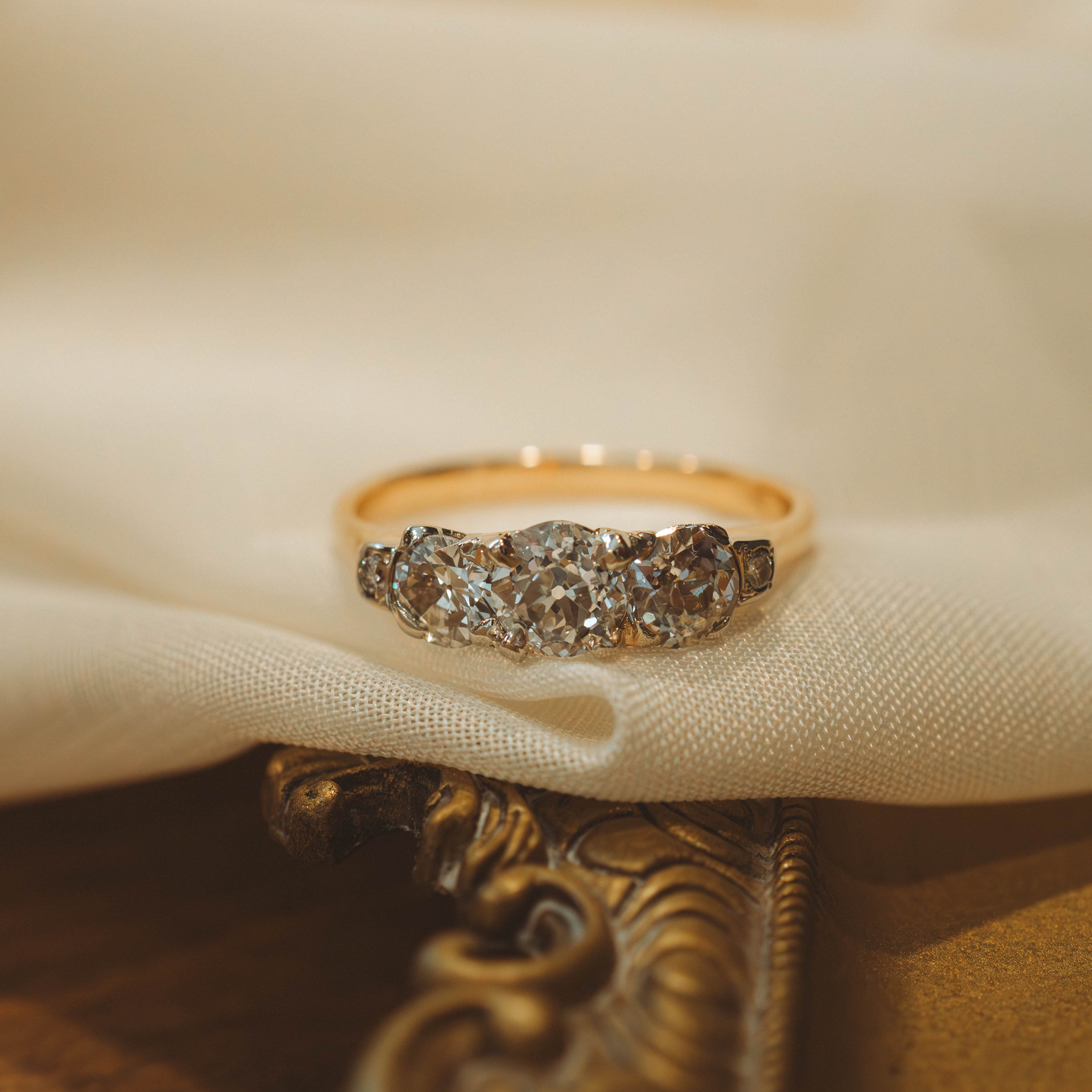 A gold ring with 3 diamonds, resting on draped cream-colored fabric over an ornate gold surface.