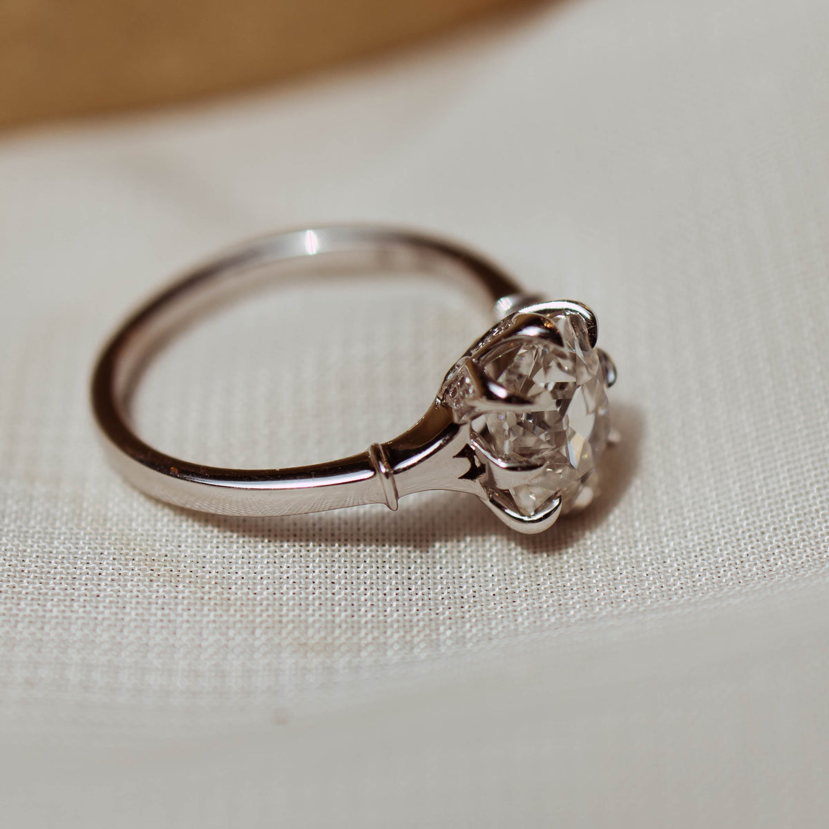 A silver-toned solitaire ring with an old mine cut diamond e in a prong setting, resting on a textured white fabric surface with a warm, blurred gold-toned background.