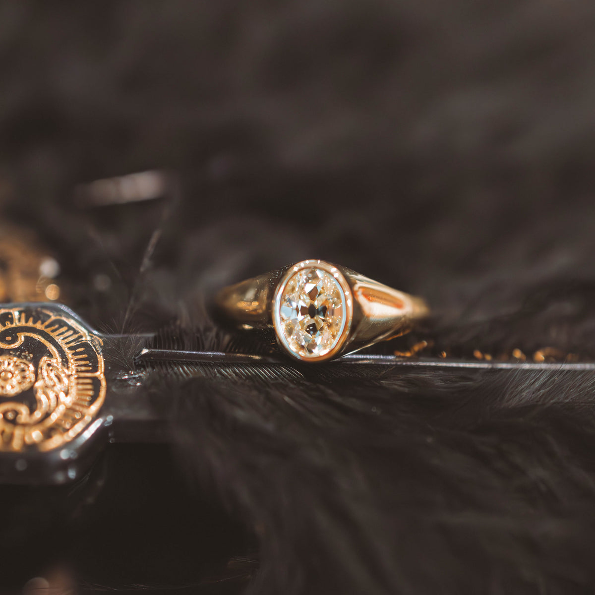 A gold signet ring with an oval-cut clear gemstone, resting on a black feather next to a gold seal, set against a dark, fluffy black background.