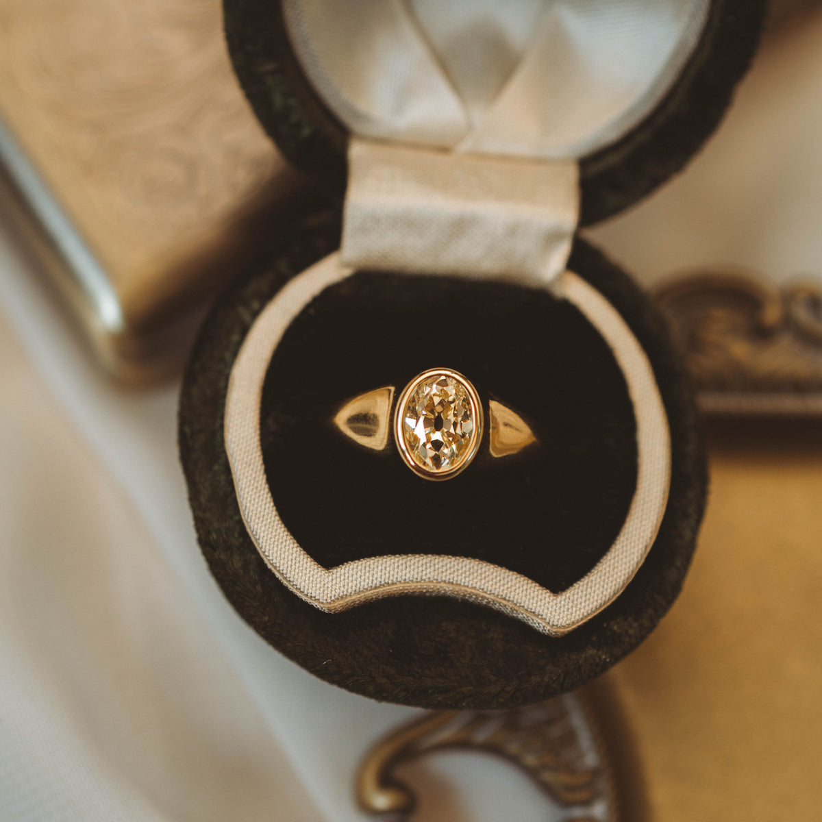 A gold ring with an oval yellow diamond set in a black velvet ring box with a cream satin lining, photographed on a warm beige surface.