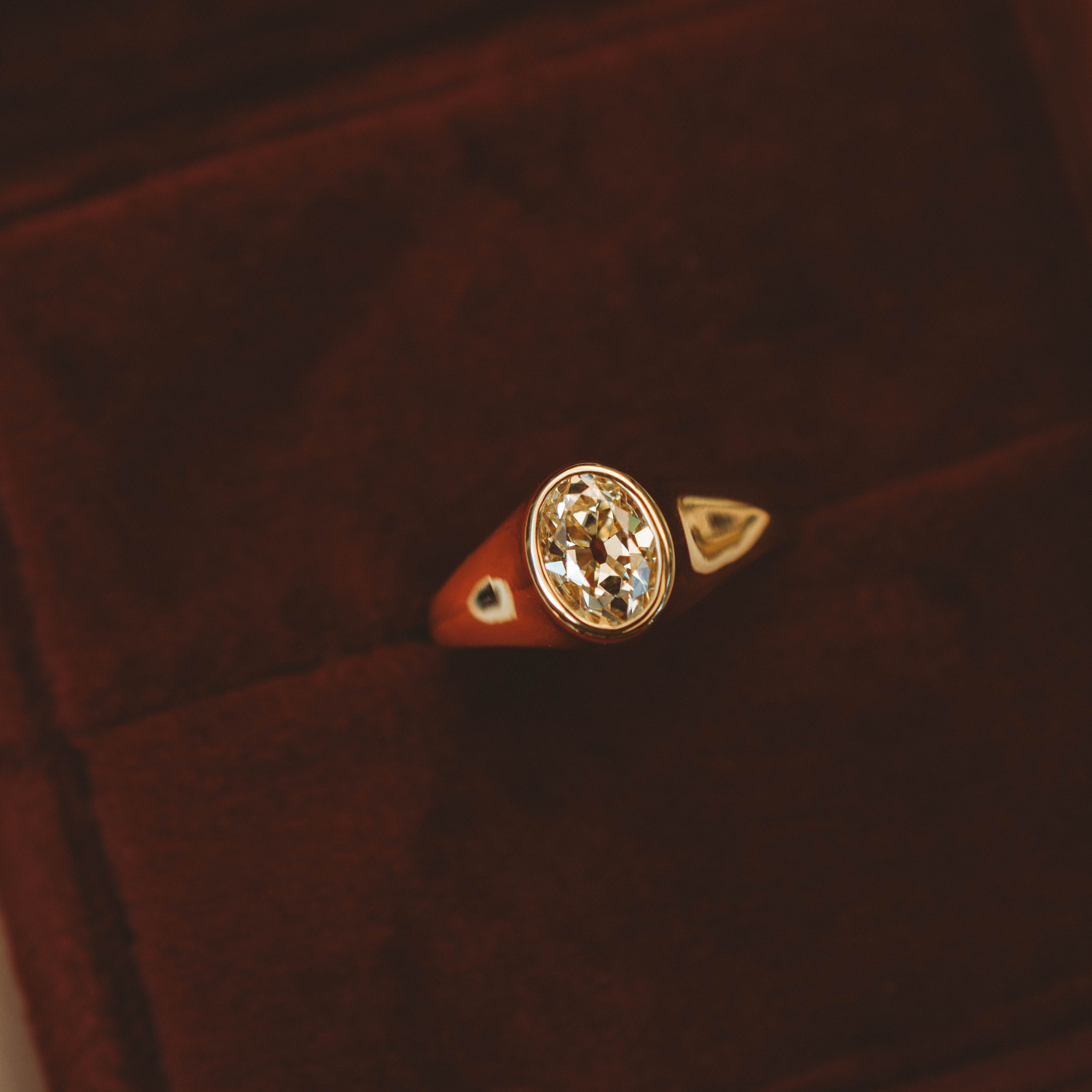 A gold signet-style ring with an oval diamond displayed in a dark brown velvet ring box.