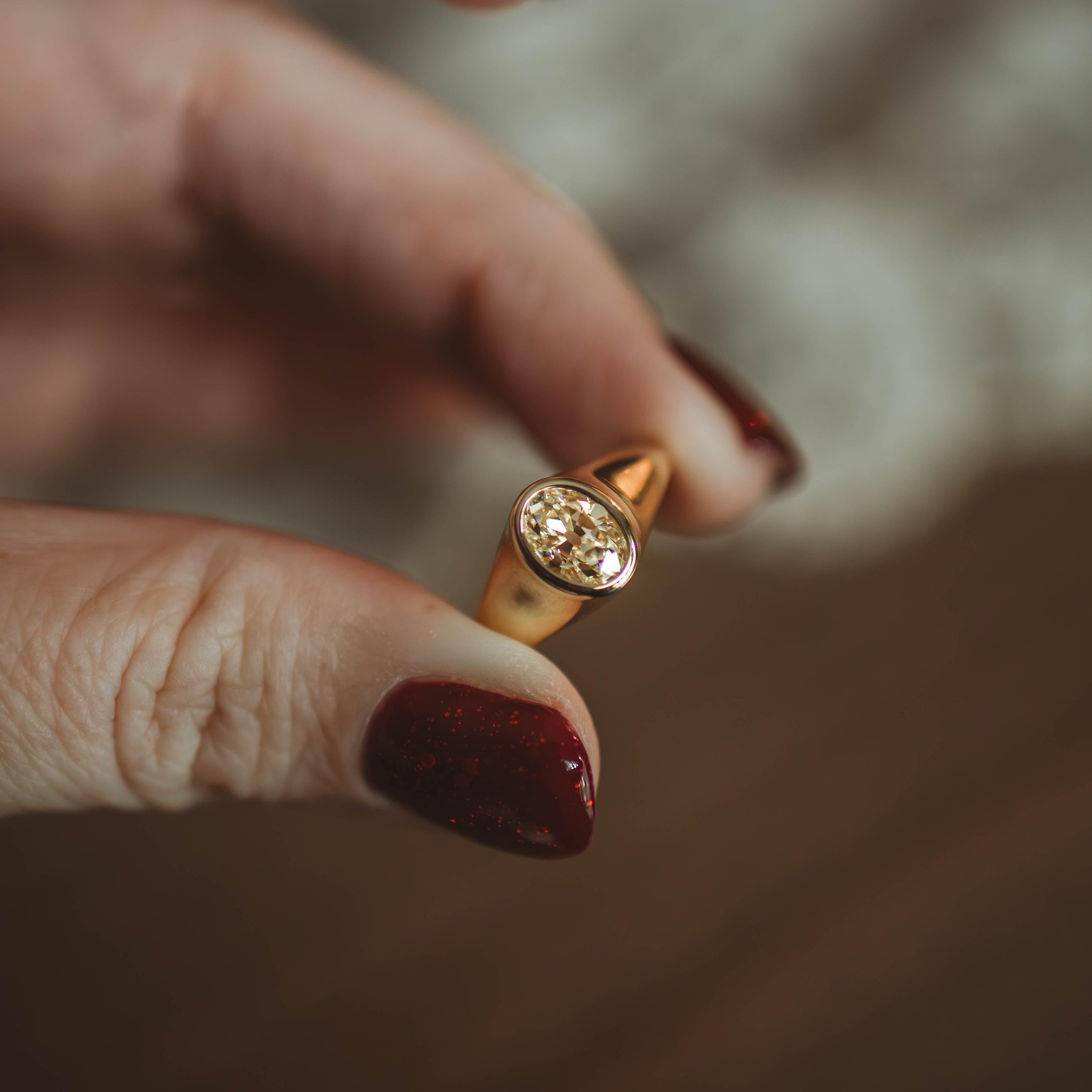 A close-up of a hand with dark red glitter nail polish holding a gold signet ring with a round yellow diamond against a warm, blurred background.